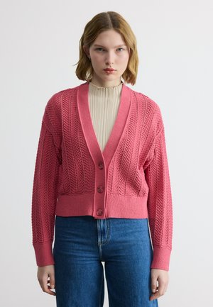 Jonge vrouw die een roze gebreid vest met knopen draagt over een beige coltrui en een blauwe spijkerbroek tegen een witte achtergrond.