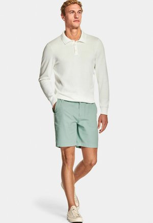 Man loopt met een witte poloshirt met lange mouwen, mintgroene shorts en ecru sneakers tegen een effen witte achtergrond.