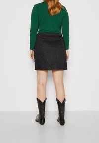 Jupe noire avec une texture légèrement scintillante, associée à un haut à manches longues vert. Présente une coupe arrivant aux genoux et des bottes de cowboy noires avec des accents détaillés.