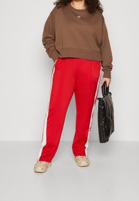 Person som bär en brun sweatshirt och röda träningbyxor med vita ränder, håller en svart vadderad handväska, står på ett grått golv.