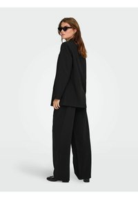Sort oversized blazer med matchende brede bukser. Båret med sorte solbriller, der har lige linjer og en struktureret stoffinish.