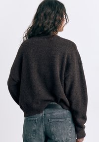 Donna con capelli lunghi e ricci che indossa un maglione largo marrone scuro e jeans a vita alta sbiaditi, vista di spalle su uno sfondo semplice.