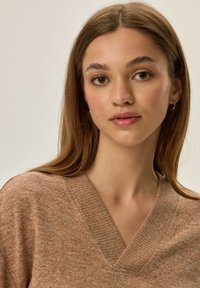 Brązowy sweter w dekolcie w kształcie litery V o teksturowanym wykończeniu, z luźnym krojem i subtelnymi, melanzowymi wzorami. Miękka tkanina delikatnie opada.