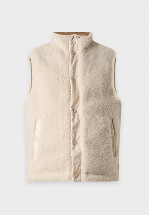 Homme portant une veste polaire crème sur une chemise noire à manches longues, un jean foncé et des baskets beige clair, debout avec une main dans la poche.