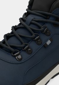 Navy hiking shoe with textured synthetic upper, rounded toe, and black hardware. Features contrasting laces and a sturdy sole for grip.