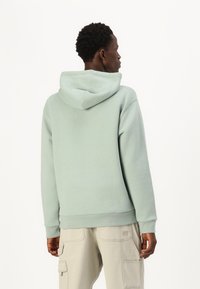 Hanorac cu glugă dintr-un material moale de culoare verde mentă, cu o croială lejeră. Include o buzunare tip kangaroo și manșete și tiv cu model ch rib. Asortat cu pantaloni cargo deschiși.