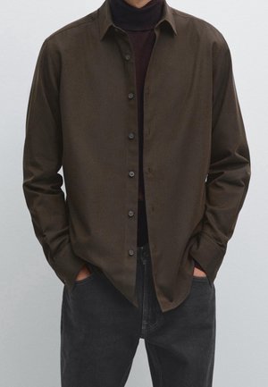 Homme portant une chemise marron à boutons sur un col roulé foncé, les mains dans les poches, associé à un jean noir sur un fond clair uni.