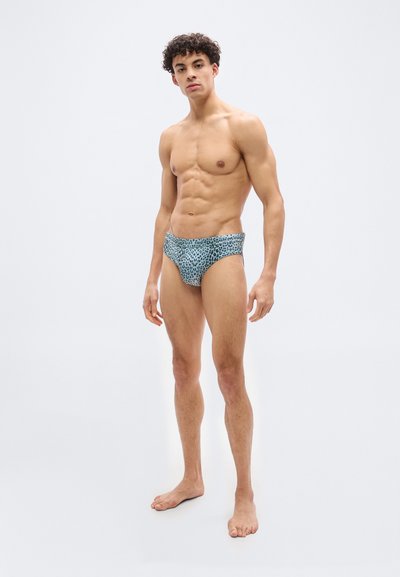 Μυώδης άνδρας όρθιος, ξυπόλητος, φοράει μπλε εσώρουχα με λεοπάρ μοτίβο, μπροστά σε απλό φωτεινό φόντο, κοιτάζοντας προς τα εμπρός.