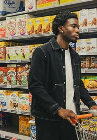 Veste en denim noire avec des coutures blanches, portée sur une chemise blanche. Un chariot de magasin à la main, entouré de boîtes de céréales colorées. Cadre de vente au détail.