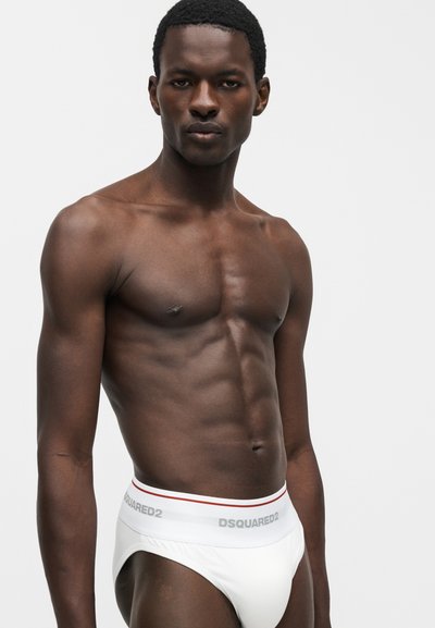 Μυώδης άνδρας με σκουρόχρωμο δέρμα που φοράει λευκά εσώρουχα DSQUARED2 στέκεται μπροστά σε απλό ανοιχτό γκρι φόντο.