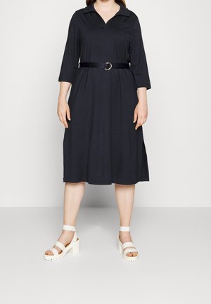 Kvinde iført en sort midi-kjole med 3/4-ærmer, et sort bælte og hvide hælede sandaler, stående mod en ensfarvet lys baggrund.