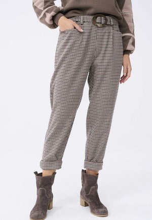 Person trägt karierte Hose mit hoher Taille, breitem Gürtel und braunen Wildleder-Stiefeletten, Hand in der Tasche vor einfarbigem Hintergrund.