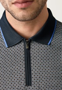 Rövid ujjú póló fekete geometriai mintával, cipzáras nyakkal, texturált gallérral és kék kontrasztos részletekkel.