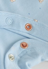 Material de bumbac albastru deschis cu modele mici de ursuleți, având trei capse cu clichet: una aur roz, una albastru deschis și una argintie.