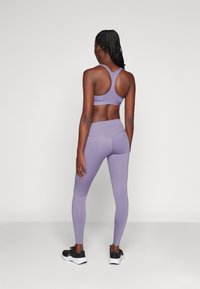 Lila sportmelltartó és hozzáillő leggings rugalmas anyagból. Racerback stílusú, testhez simuló fazonú és varrás nélküli részletekkel rendelkezik. Fekete sportcipők.