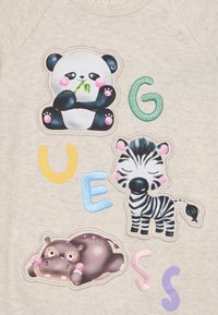 Un tissu gris clair présente des appliqués d'animaux colorés : un panda, un zèbre et un hippopotame, accompagnés de lettres multicolores en gras formant le mot "GUESS."