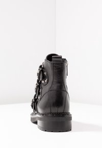 Černá kožená kotníková bota s kovovými akcenty, dekorativními smyčkami a texturovaným povrchem. Obsahuje kulatou špičku a silnou podrážku.