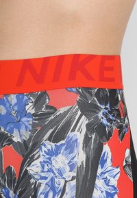 Cintura elastica rossa con il testo "NIKE", abbinata a leggings aderenti con un motivo floreale nero e blu su uno sfondo rosso vivace.