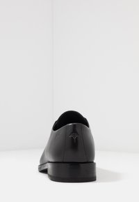 Scarpa da uomo in pelle nera con una superficie liscia, forma arrotondata e tacco basso incollato. Presenta un piccolo logo impresso sul retro.