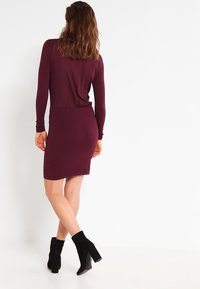 Robe bourgogne à manches longues et jupe texturée, à la coupe ajustée, associée à des bottines noires et une silhouette épurée.