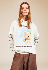 Biela košeľa s dlhými rukávmi a modrou grafikou medvedíka, pod ňou text "PONDELOK JU ANI NEPOZNAL", spolu s hnedými šortkami.