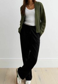 Cardigan di maglia verde oliva indossato su una canotta bianca, abbinato a pantaloni neri con coulisse e sneakers bianche con dettagli verdi.
