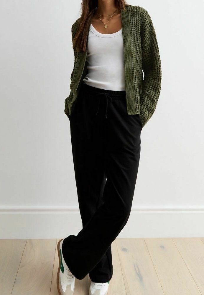 Cardigan di maglia verde oliva indossato su una canotta bianca, abbinato a pantaloni neri con coulisse e sneakers bianche con dettagli verdi.