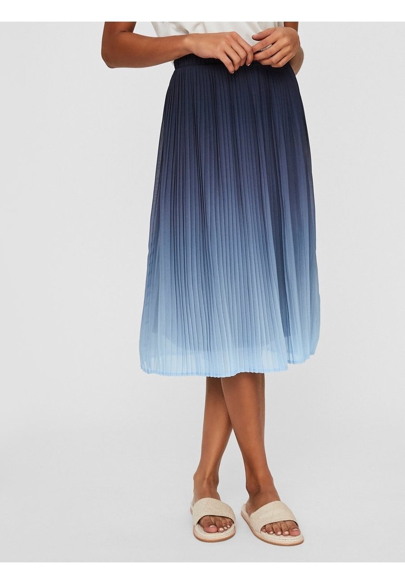 Vrouw in een blauwe ombre geplooide midi-jurk en beige slide sandalen, staande met handen op de taille op een witte achtergrond.