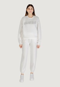Biały komplet bluzki typu sweatshirt i spodni dresowych z bawełny z błyszczącym logo "GUESS", ściągaczami przy rękawach oraz dwoma bocznymi kieszeniami. Zestawiony z białymi trampkami.