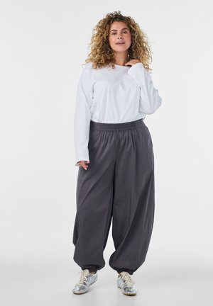 Vrouw met krullend haar, die een wit overhemd met lange mouwen, losse donkergrijze broek en zilveren sneakers draagt, staand tegen een effen achtergrond.