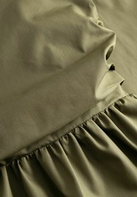 Ύφασμα ελιάς πράσινο με ραφή που συγκεντρώνει δημιουργώντας απαλά πτυχώσεις και υφή, δείχνοντας λεία και ματ λεπτομέρειες υλικού.