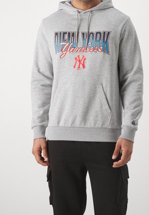 Mężczyzna noszący jasnoszarą bluzę z kapturem z napisem i logo „New York Yankees” w kolorze niebieskim i czerwonym, w połączeniu z czarnymi spodniami typu cargo.