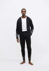 Čierna mikina na zips s kapucňou, sivými akcentmi a bielym lemom, v páre s čiernymi teplákmi s podobným lemom. Model stojí bosý na svetlom pozadí.