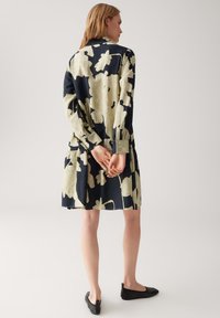 Frau steht mit hinter dem Rücken verschränkten Händen, trägt ein lockeres schwarzes Kleid mit großem beigen Blumenmuster und schwarze flache Schuhe.