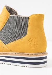 Żółty zamszowy botek sięgający do kostki z panelem z tkaniny w jodełkę, perforacjami, tłoczonym logo oraz gumową podeszwą w wielokolorowe paski.