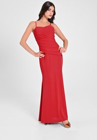 Rotes, ärmelloses Kleid mit gerafftem Oberteil, fließender Silhouette und bodenlanger Gestaltung. Kombiniert mit goldenen Akzent-Absatzschuhen. Glatter Stofftextur.