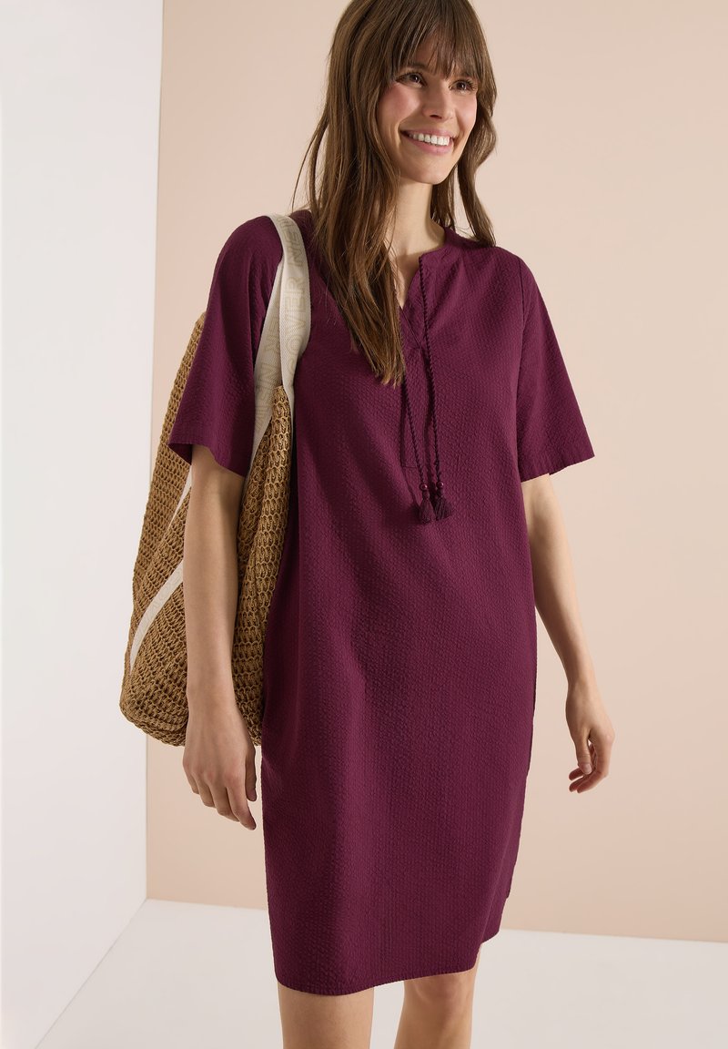 Femme souriante portant une robe violette texturée à manches courtes avec des liens à pompons, portant un grand sac à épaule tissé.