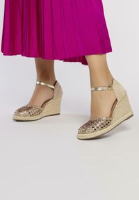 Sandalias en oro con cuña tejida, diseño de punta abierta y tira en el tobillo; adornadas con un patrón brillante en la parte frontal.