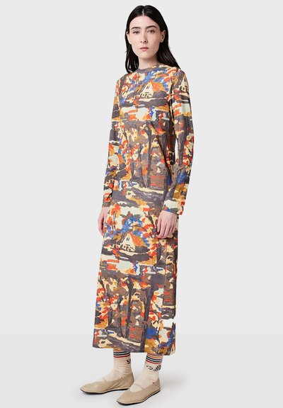 Kobieta ubrana w długą sukienkę w wielokolorowy wzór abstrakcyjny z długimi rękawami, w paskowane skarpetki i beżowe płaskie buty, stojąca na jednolitym tle.