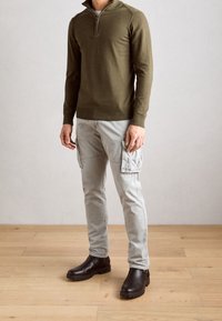 Olivgrüner Pullover mit Reißverschluss am Kragen, hellgraue Cargo-Hose mit Seitentaschen und dunkelbraune Stiefeletten auf einem Holzboden.