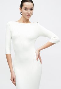 Γυναίκα που φοράει εφαρμοστό λευκό φόρεμα ριπ με μανίκια τριών τετάρτων και χρυσά κουμπιά στο μανίκι, ποζάροντας με το ένα χέρι στη μέση.