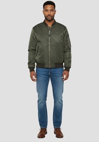 Bomberjack in donkergroen, gemaakt van gladde stof, met een ritssluiting aan de voorkant, geribde manchetten en zijzakken. Gecombineerd met een blauwe jeans en bruine schoenen.