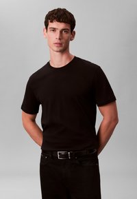 T-shirt en coton noir à manches courtes, col rond, et texture lisse, associé à un jean noir et une ceinture noire, sur un fond neutre.