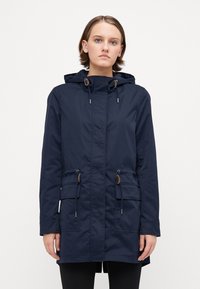 Marineblå parkajakke laget av vannavvisende stoff. Har hette, snor i midjen og to frontlommer med klaffer.