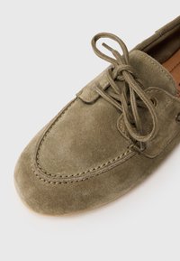 Chaussure en daim vert olive avec un bout rond, un laçage, des accents cousus et des œillets métalliques sur les côtés. Semelle en caoutchouc beige.