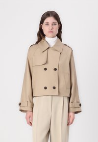 Cappotto trench cropped beige con bottoni doppi, colletto ampio e polsini arrotolati, realizzato in tessuto liscio. Tasca laterale verticale visibile.