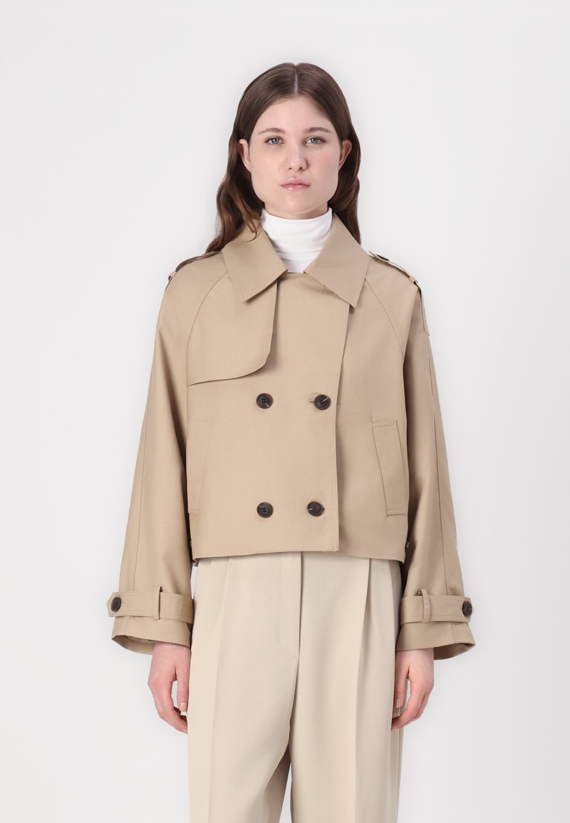 Cappotto trench cropped beige con bottoni doppi, colletto ampio e polsini arrotolati, realizzato in tessuto liscio. Tasca laterale verticale visibile.