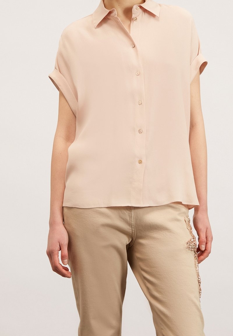 Blouse rose clair à manches courtes en tissu lisse, avec un devant boutonné et un col doux. Associée à un pantalon beige orné d'embellissements.