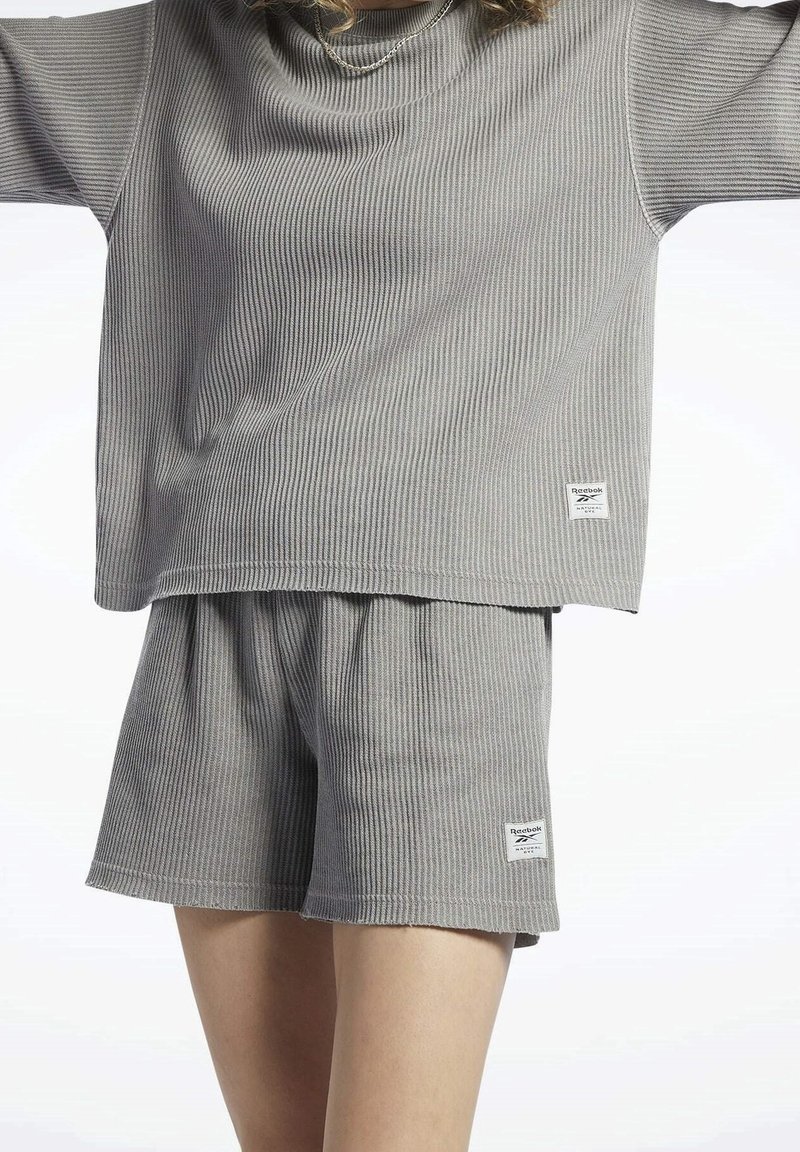 Šedá žebrovaná relaxační souprava obsahující top s krátkým rukávem a šortky. Materiál má měkkou strukturu. Obsahuje malou značkovou etiketu na každém kusu.