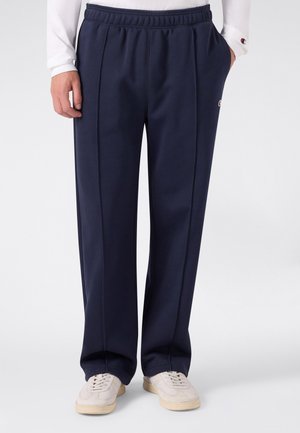Person trägt marineblaue gerade geschnittene Hose mit seitlichen Taschen und weißen Sneakers, steht vor einem schlichten hellen Hintergrund.